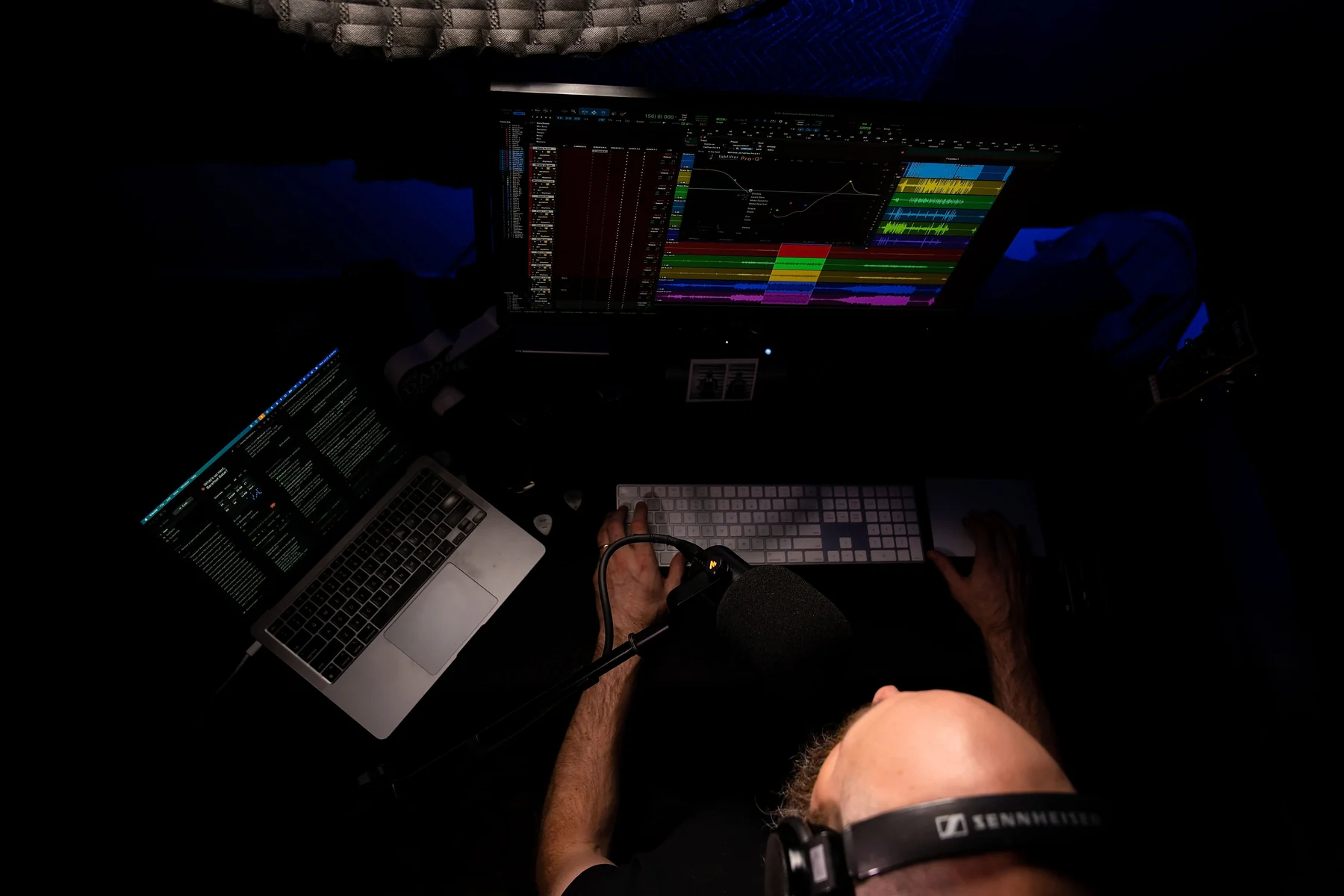 Overhead view of David Glenn's desk in the dark: a Pro Tools session with a FabFilter Pro-Q curve open on the monitor, a laptop of notes to the left, hands on the keyboard, and the Sennheiser headband across the bottom of frame